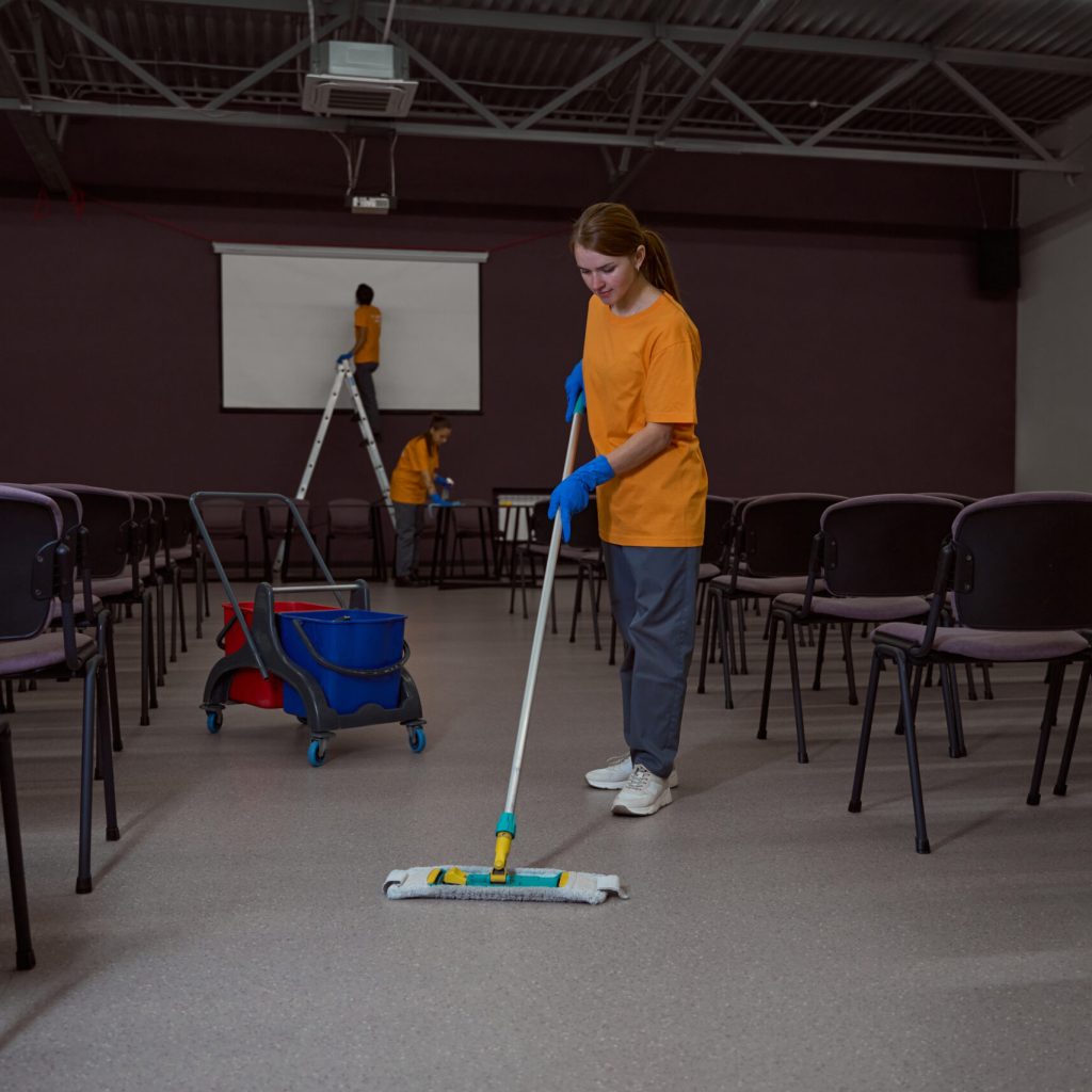 Professional janitor washing the floor with a mop while her collegues cleaning furniture in the background